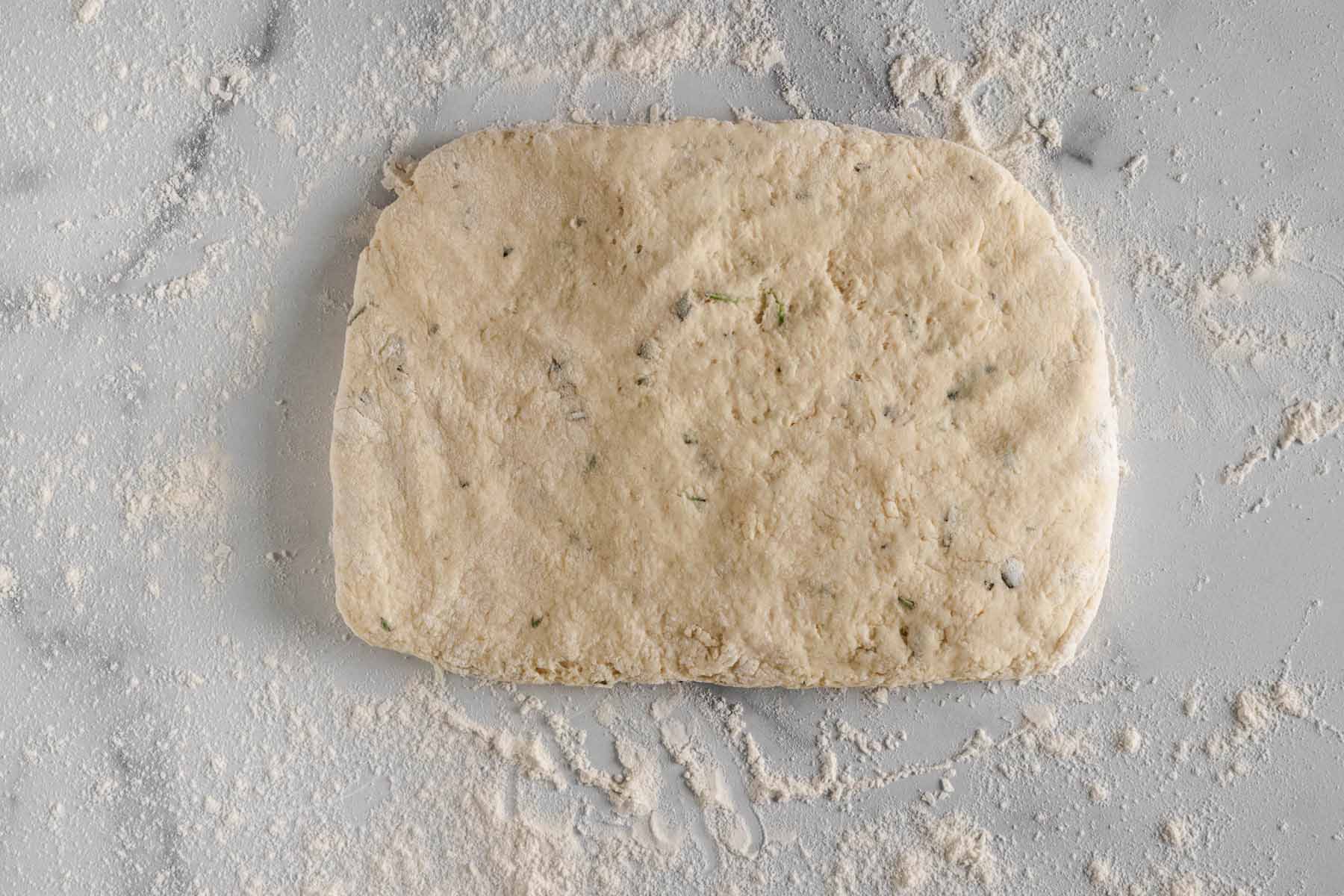 Rosemary biscuit dough shaped into a rectangle on a floured marble surface.