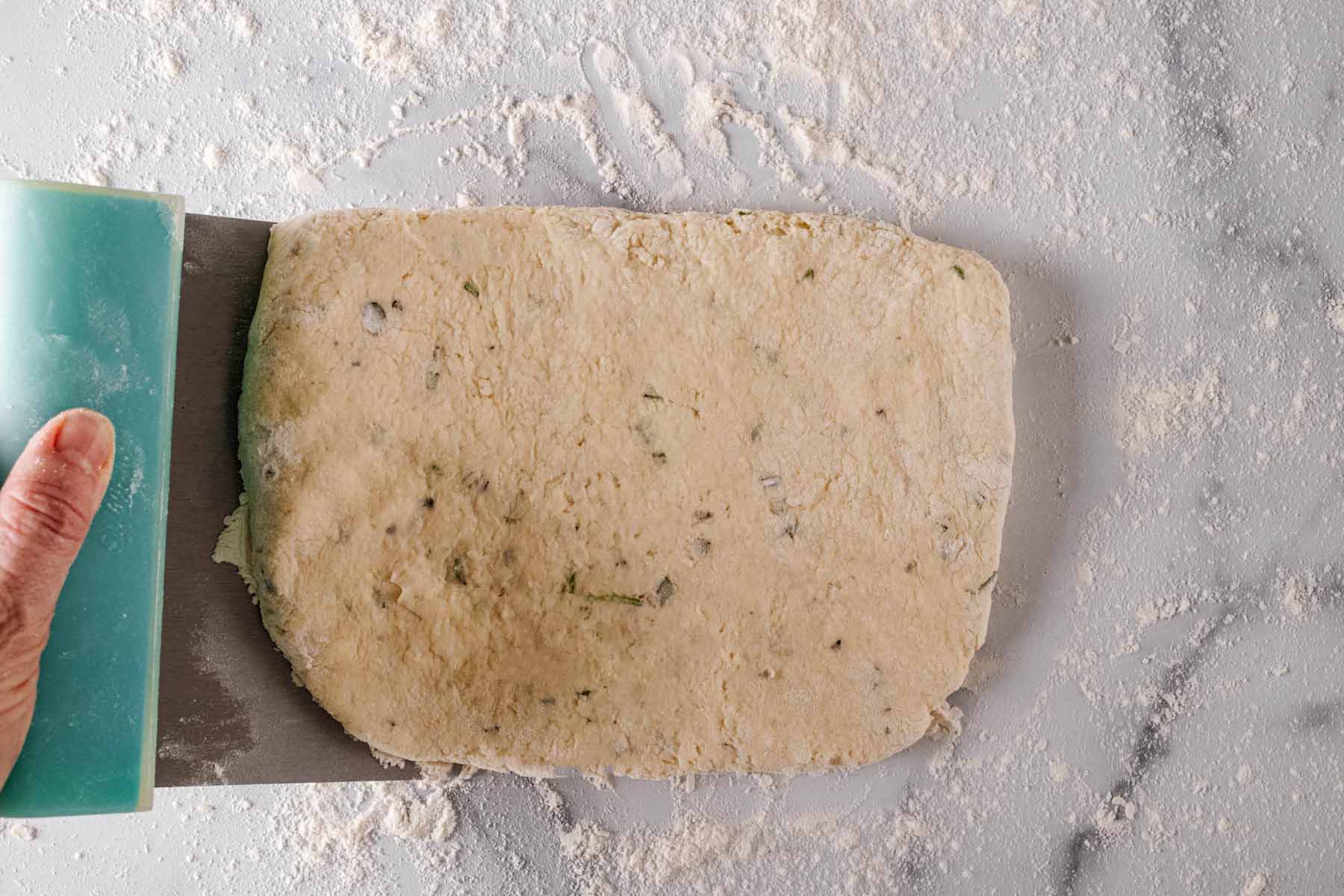 Biscuit dough being picked up on one side with a bench/dough scraper.
