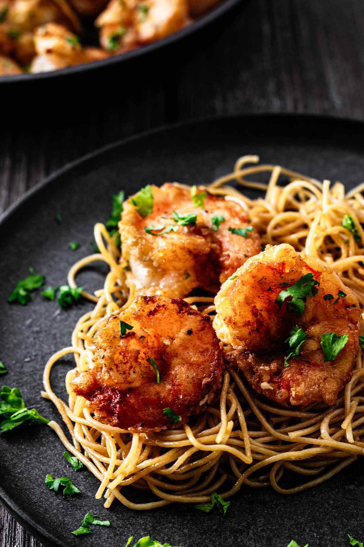 Three cooked shrimp on a bed of cooked thin spaghetti garnished with chopped parsley on a dark plate.