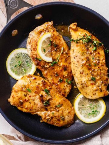 Overhead close up of three chicken limone cutlets topped with fresh thyme leaves in a skillet with sliced lemons