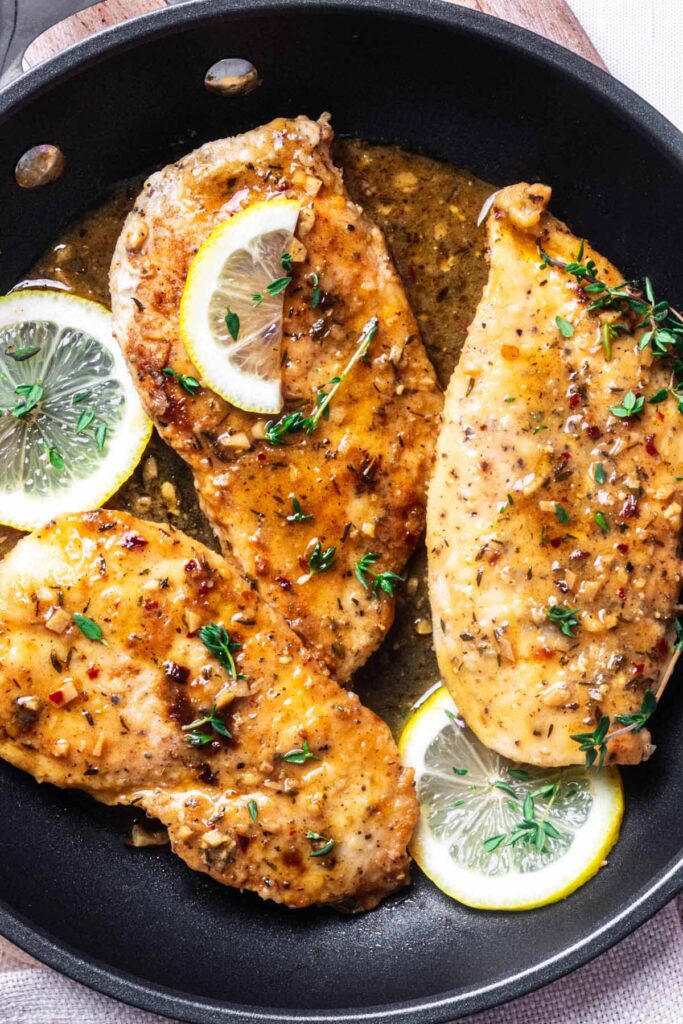 Overhe4ad close up of three chicken limone cutlets topped with fresh thyme leaves in a skillet with sliced lemons