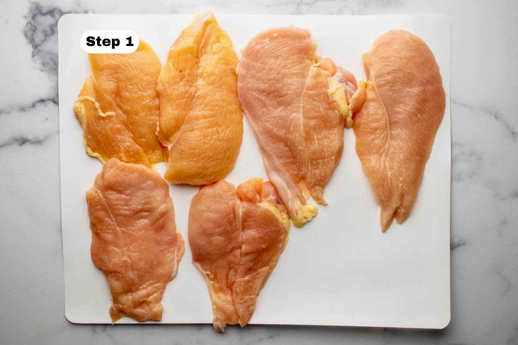 Six chicken breast cutlets on a white cutting mat.