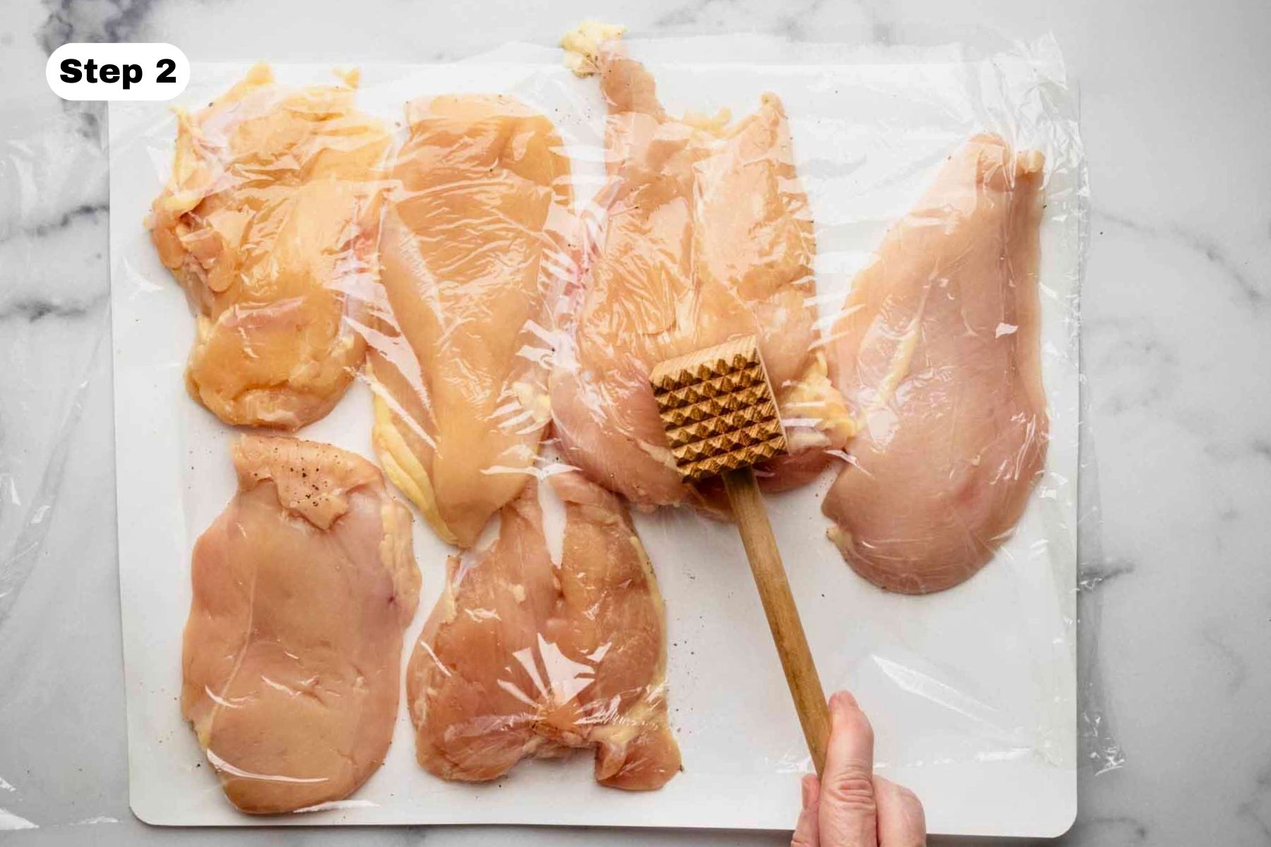 Six chicken breast cutlets covered in plastic wrap being pounded with a wooden meat tenderizer
