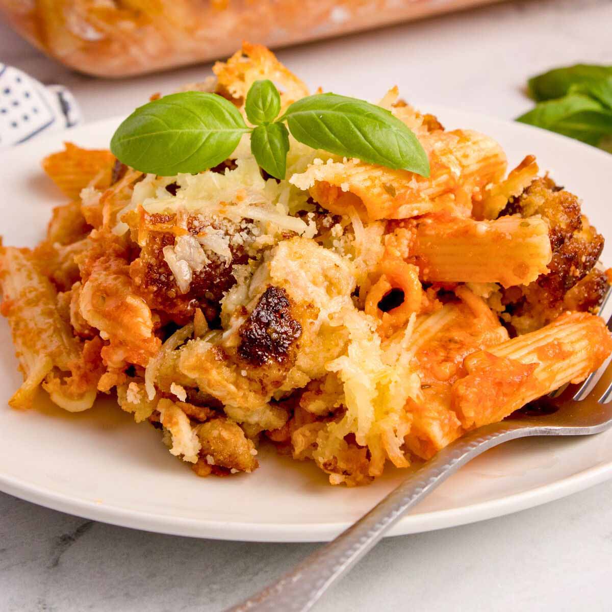 Serving of chicken Parmesan casserole topped with whole basil leaves on a white plate with a fork.