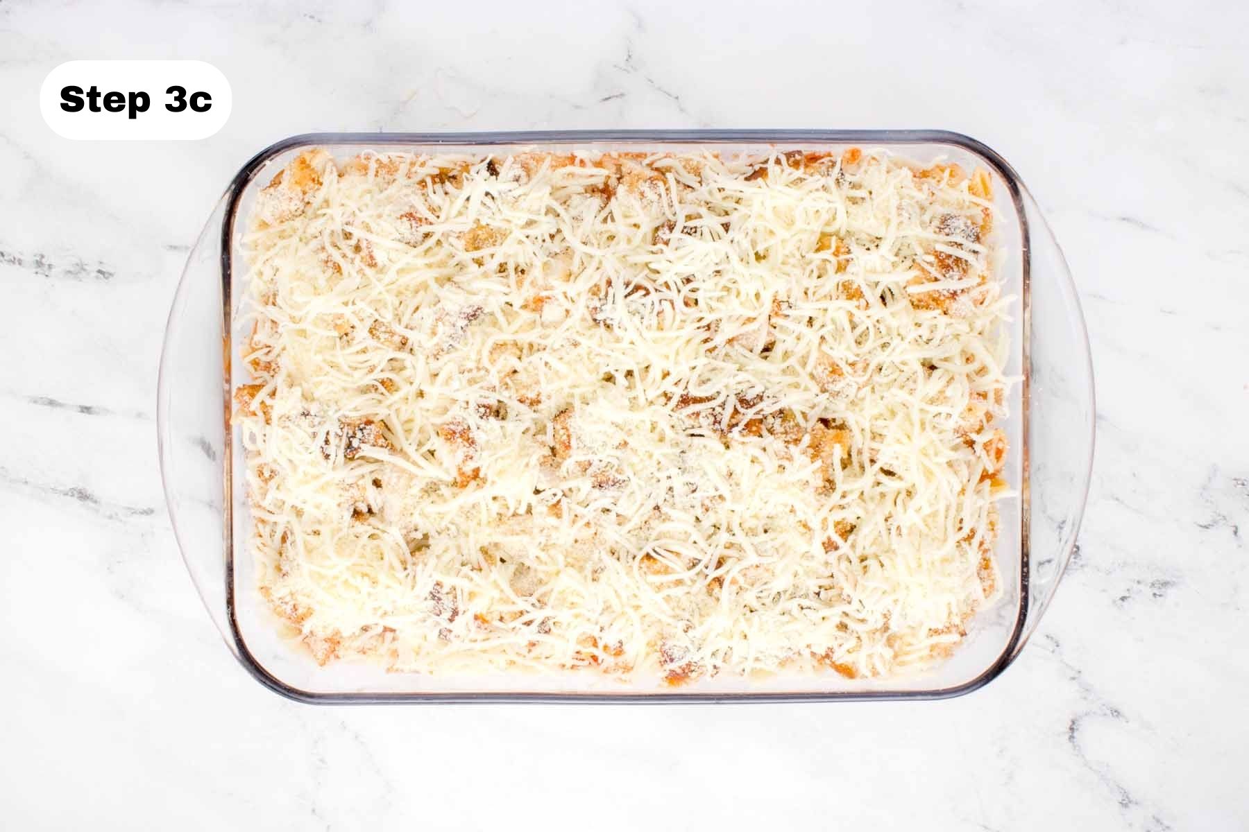 Shredded mozzarella and Parmesan layered on top of cooked chicken thigh pieces in a glass baking dish.