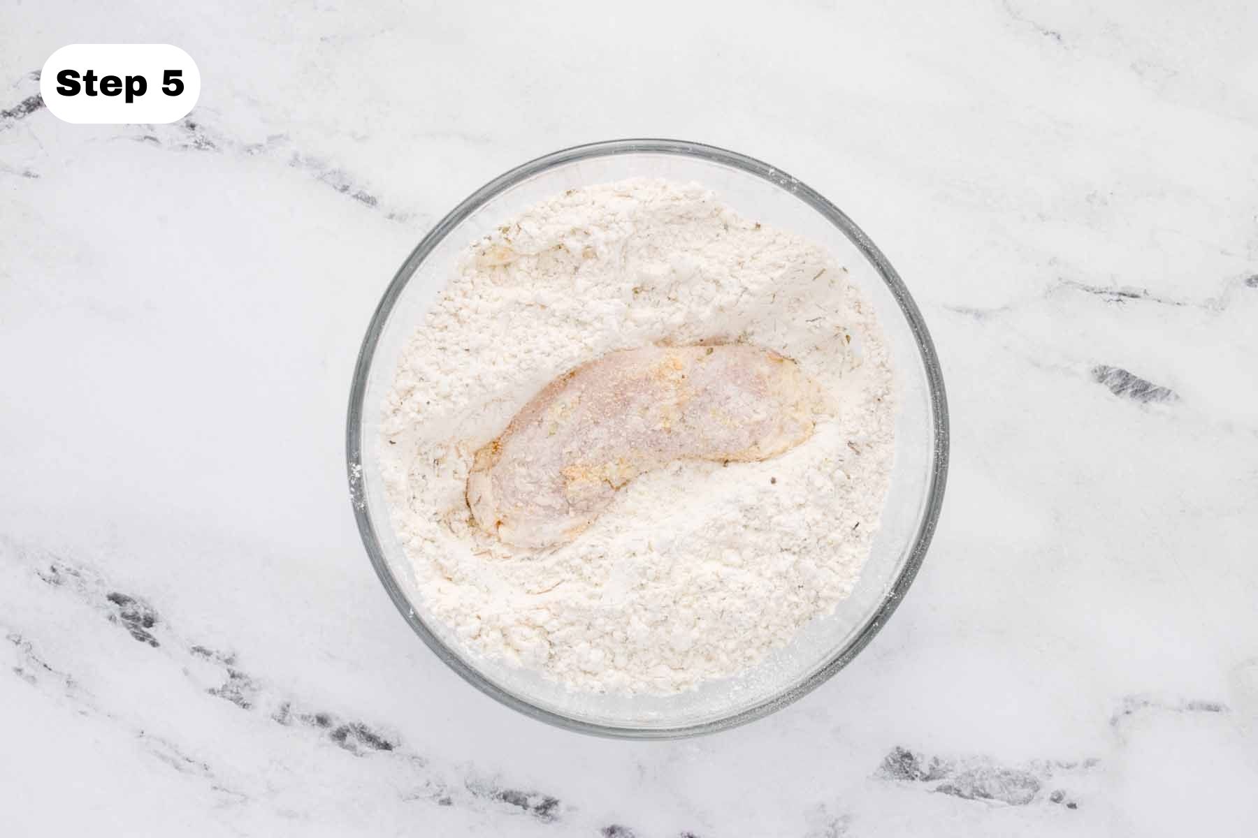 Chicken thigh in flour mixture in a glass bowl.