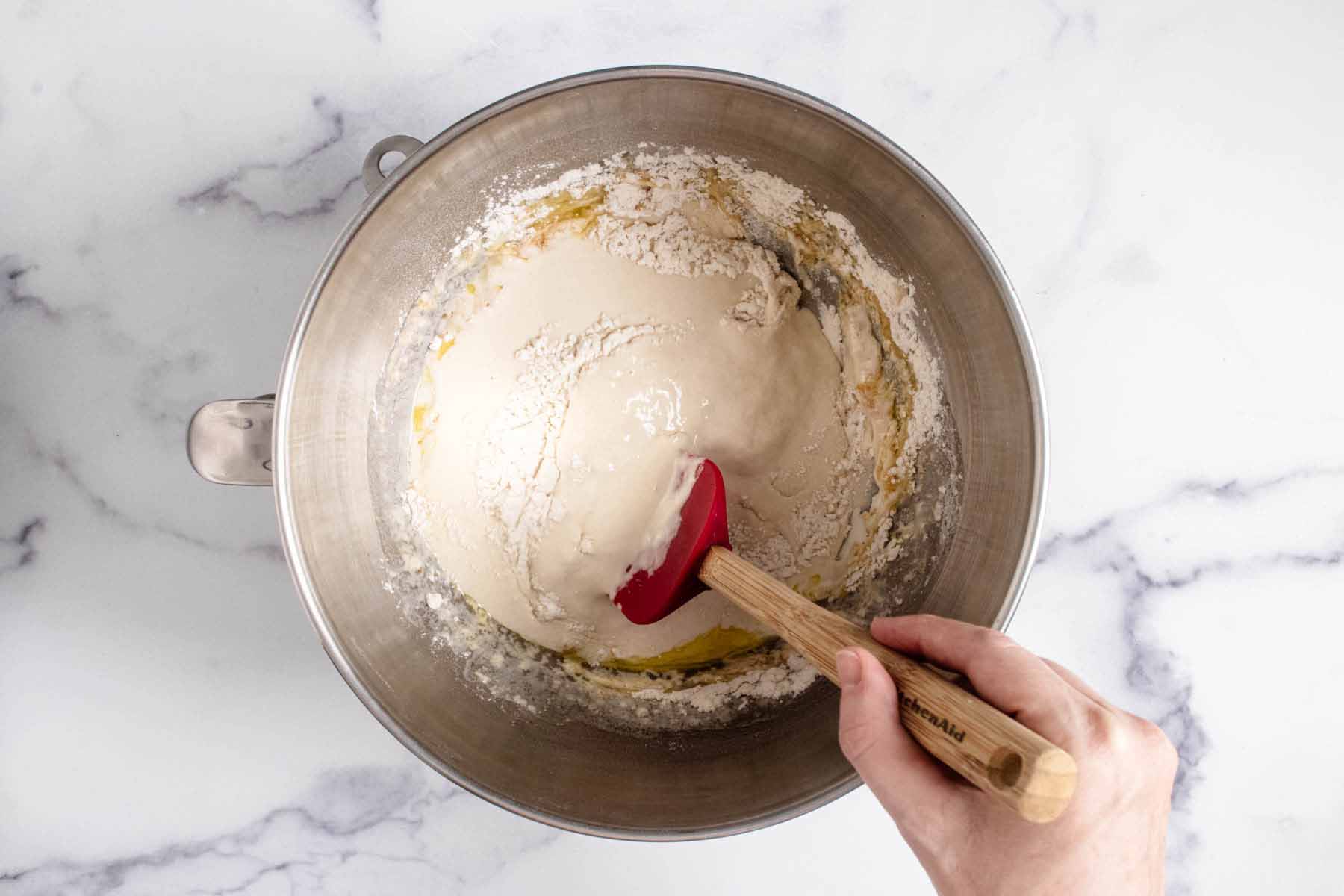 Pizza dough ingredients in a mixing bowl being combined with a rubber spatula