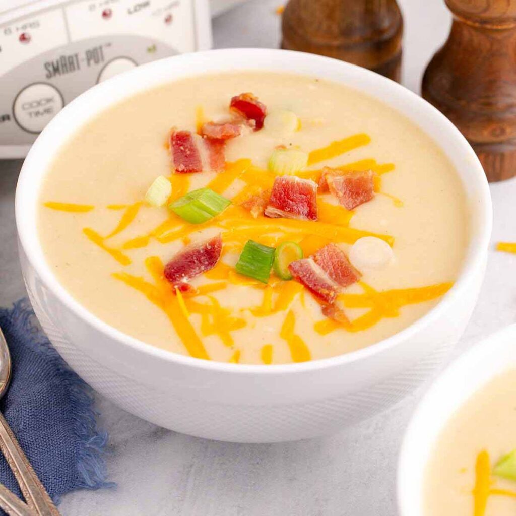 Potato soup topped with bacon bits, grated cheddar cheese, and sliced green onions in a white bowl.