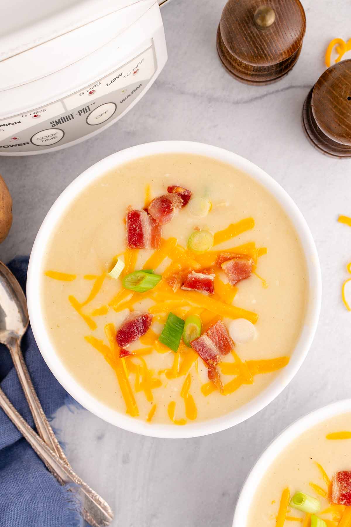 Overhead close up of slow cooker potato soup topped with bacon bits, grated cheddar cheese, and sliced green onion in a white soup bowl.