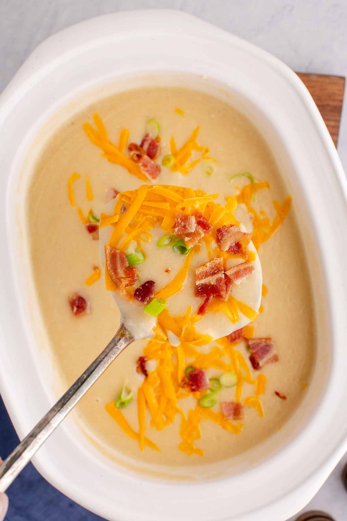 Overhead view of a ladle filled with potato soup topped with bacon bits, grated cheddar cheese, and sliced green onions over a white slow cooker filled with soup.