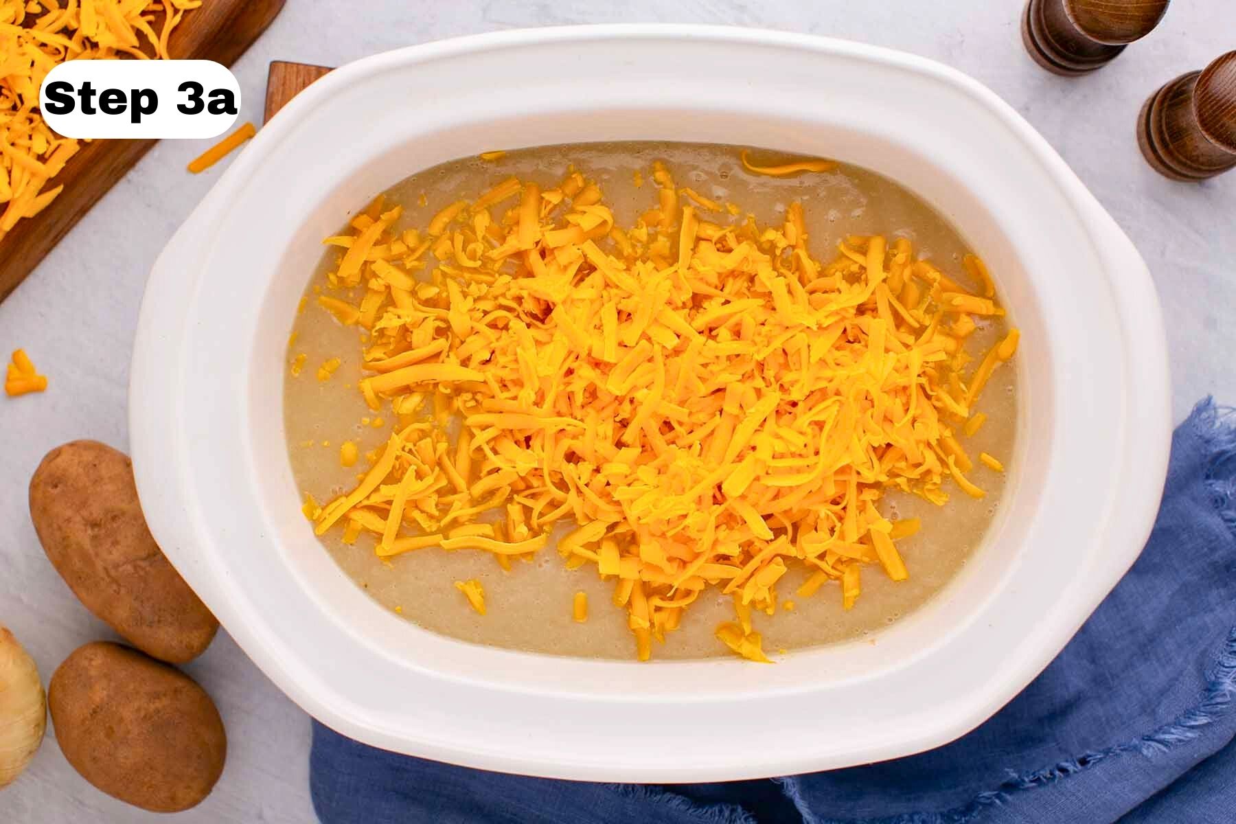 Grated cheddar cheese sprinkled over cooked soup in a large white slow cooker.