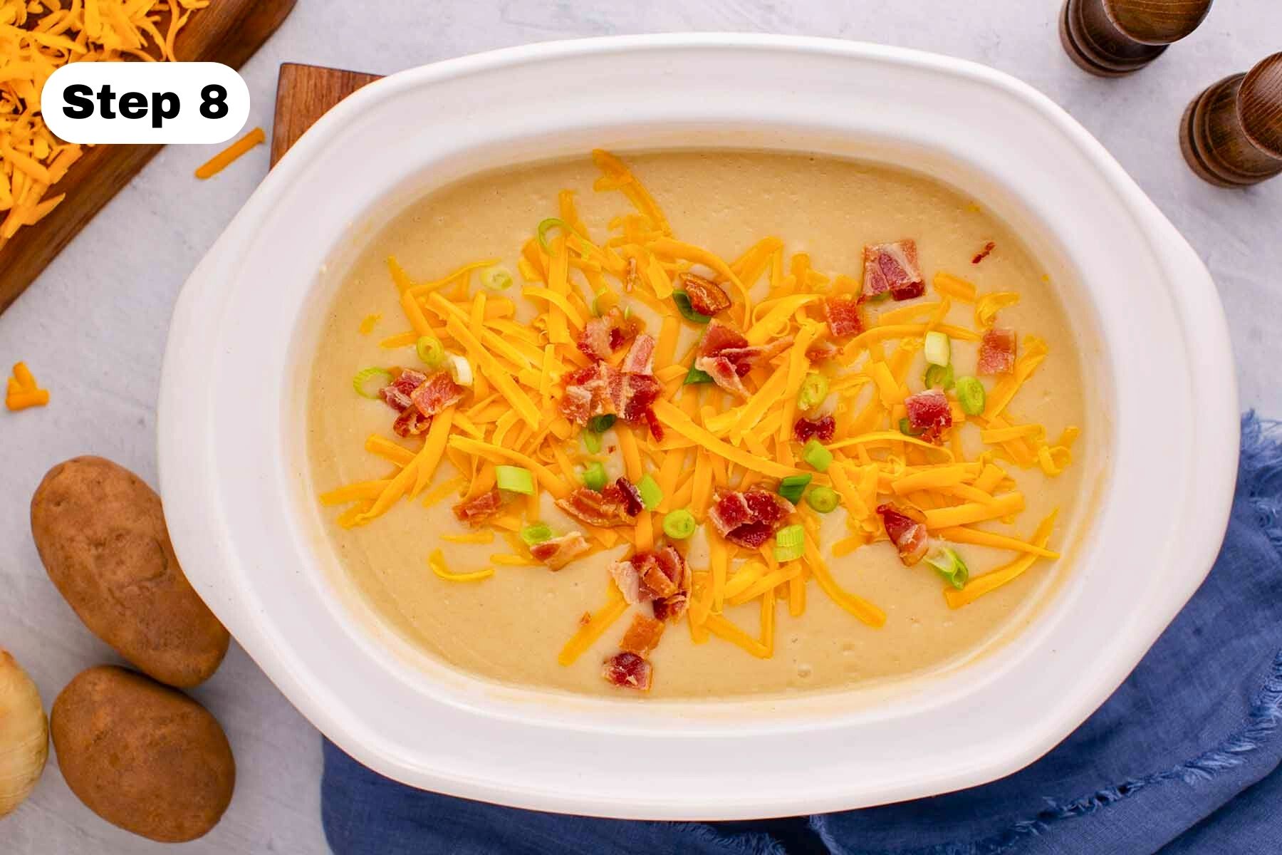 Cooked potato soup topped with bacon bits, grated cheddar cheese and chopped green onions in a large white slow cooker.