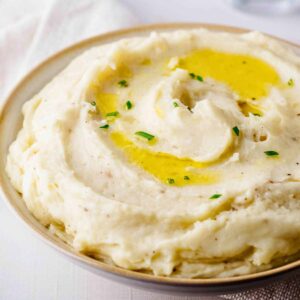 Close up of of truffle mashed potatoes garnished with olive oil and chopped chives in a light bowl with beige trim.