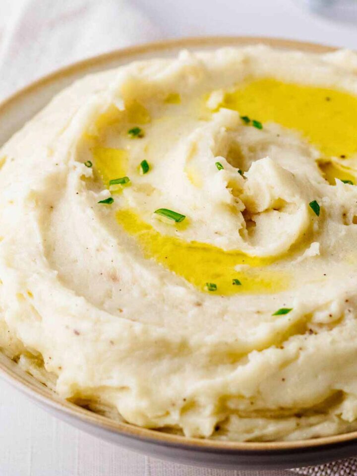 Close up of of truffle mashed potatoes garnished with olive oil and chopped chives in a light bowl with beige trim.