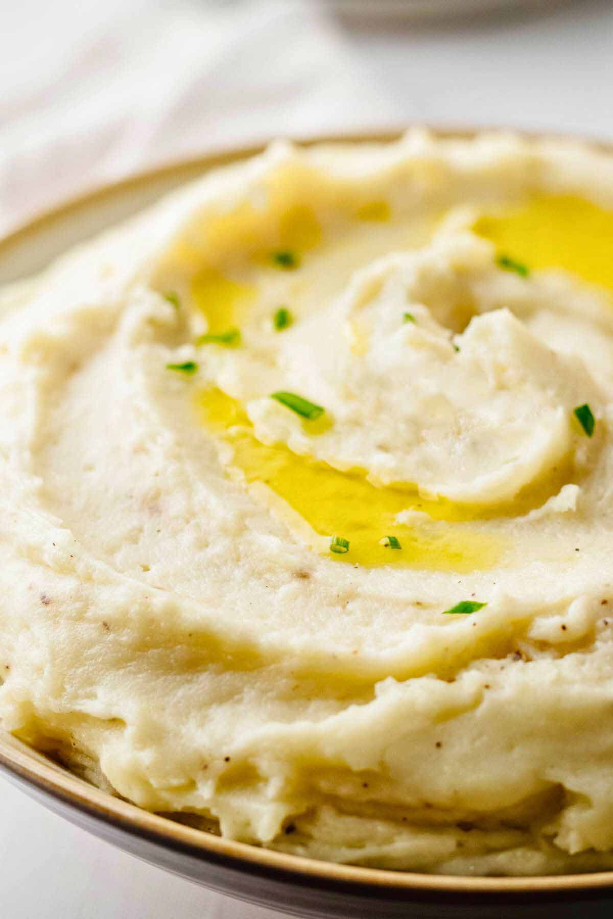 Close up of of truffle mashed potatoes garnished with olive oil and chopped chives in a light bowl with beige trim.