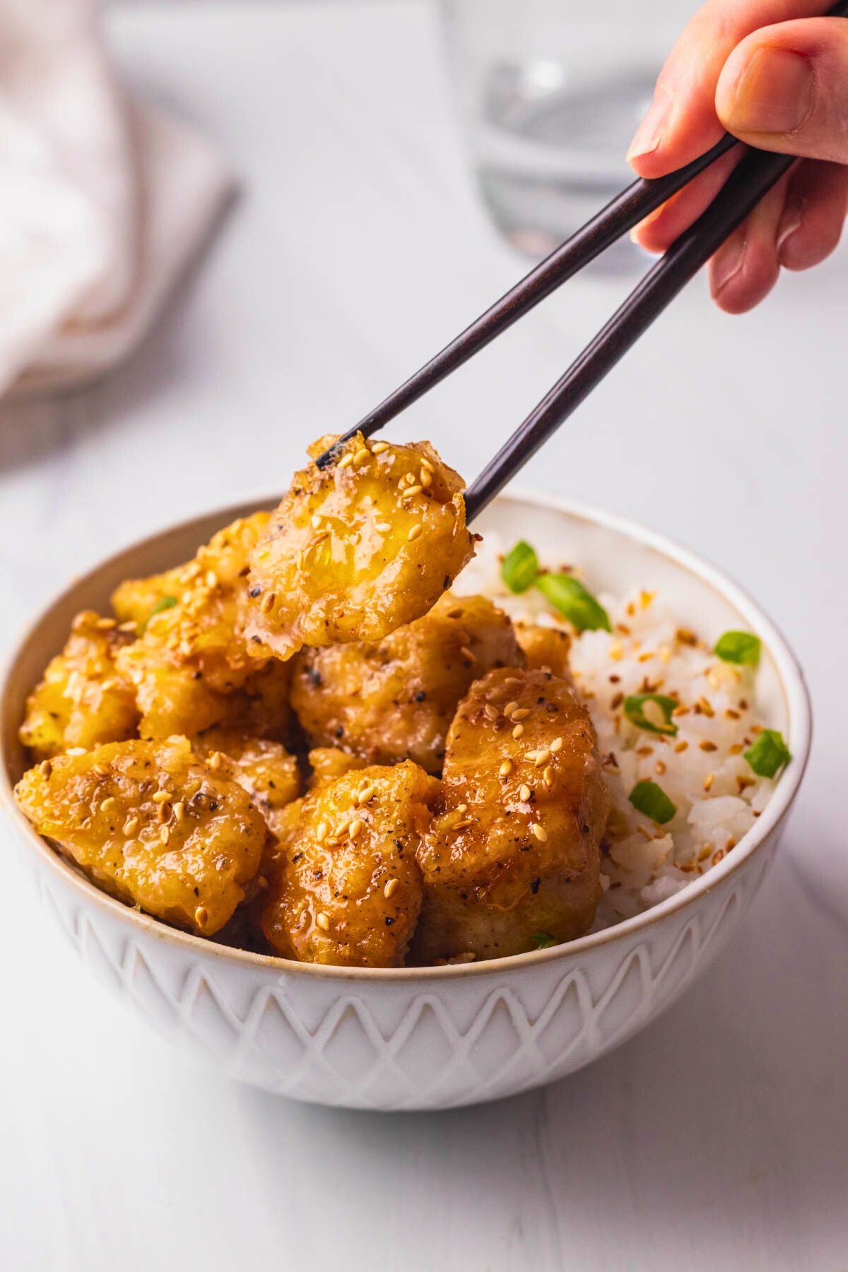 Close up of a serving of orange chicken in a white bowl and picking up a piece of chicken with chopsticks