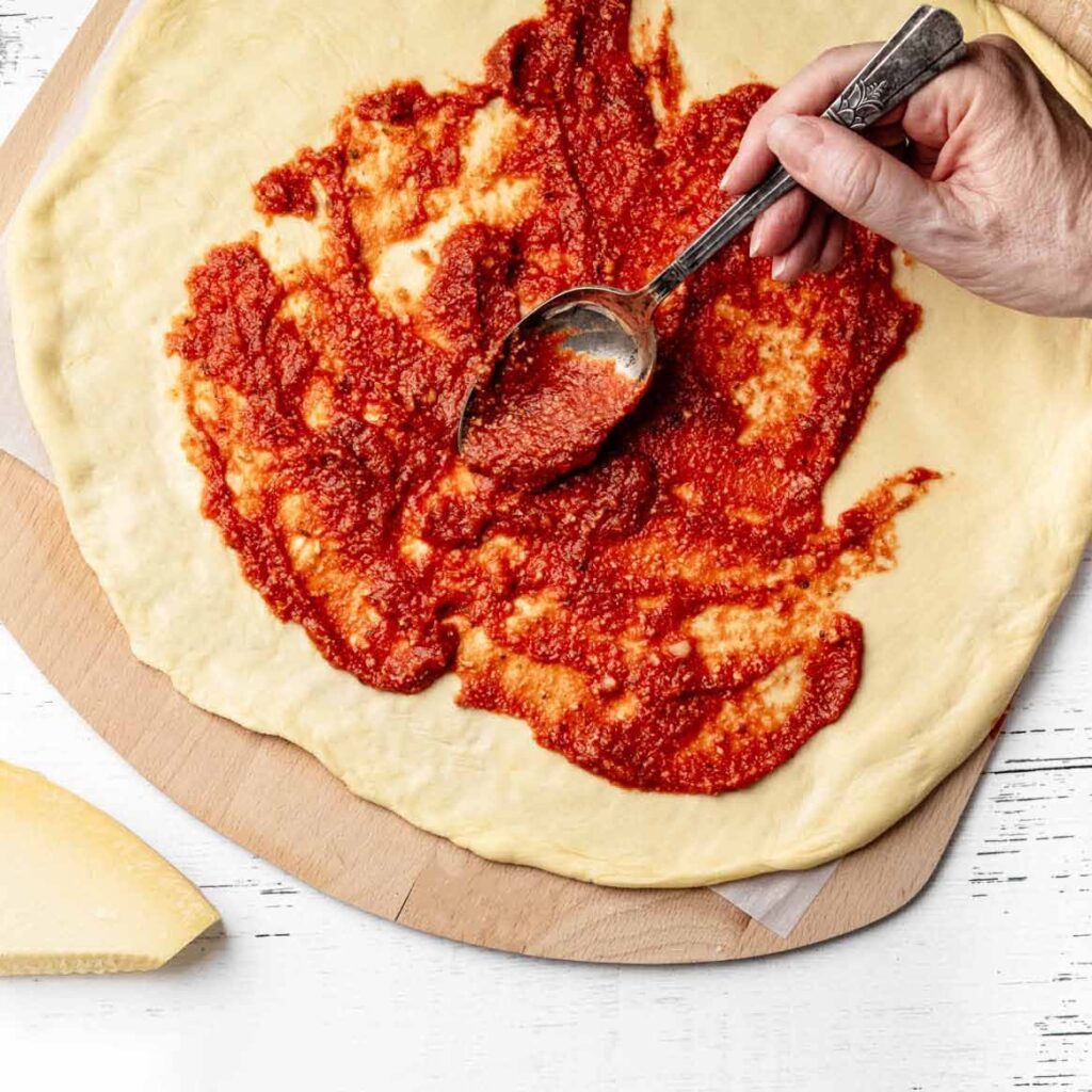 Pizza sauce being spread over raw pizza crust with a spoon