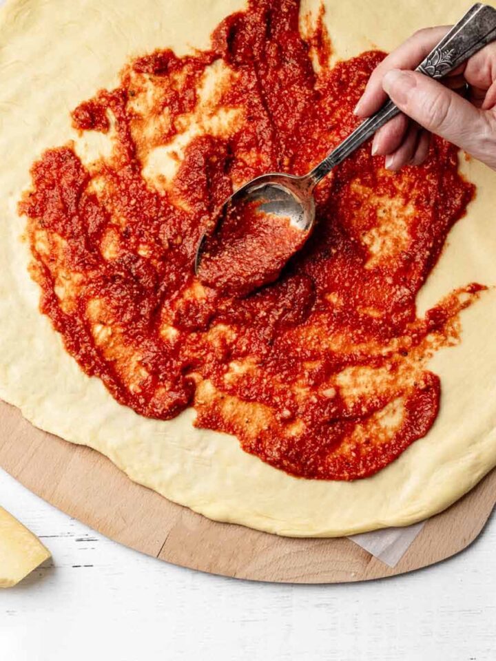 Pizza sauce being spread over raw pizza crust with a spoon