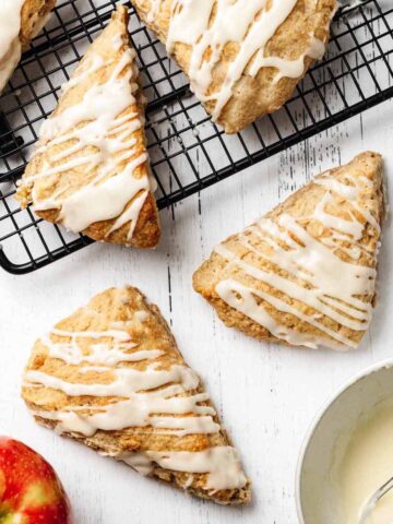 Overhead view of glazed apple scones on a white wooden surface and cooling on a wire rack.