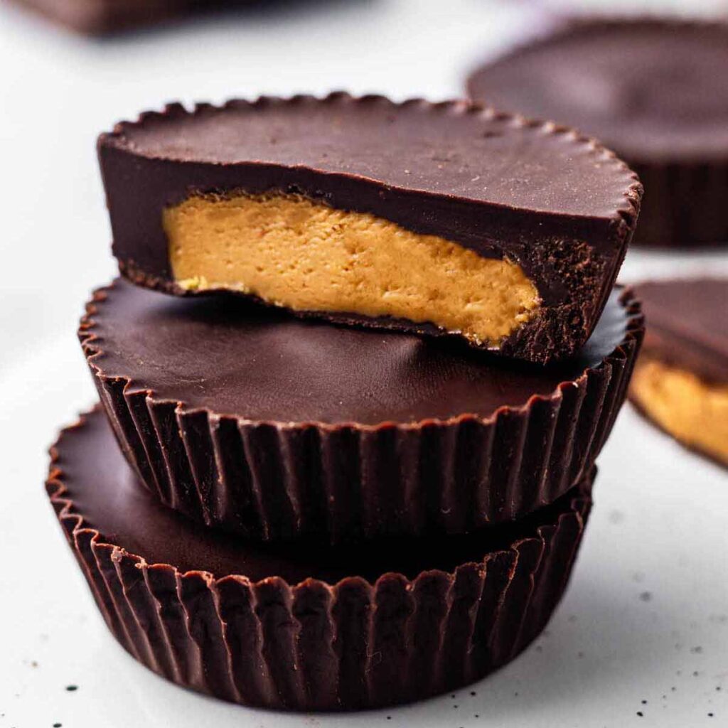 Three peanut butter cups stacked on a white surface. The top cup is cut in half to expose the creamy filling.