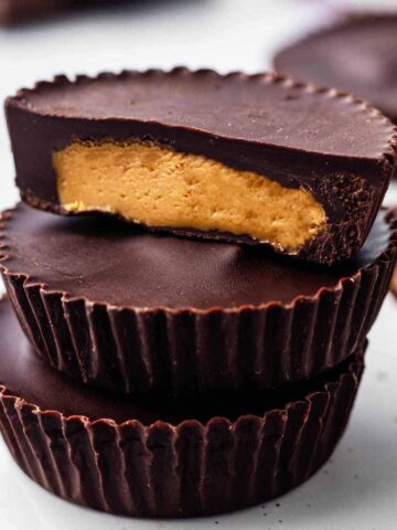 Three peanut butter cups stacked on a white surface. The top cup is cut in half to expose the creamy filling.