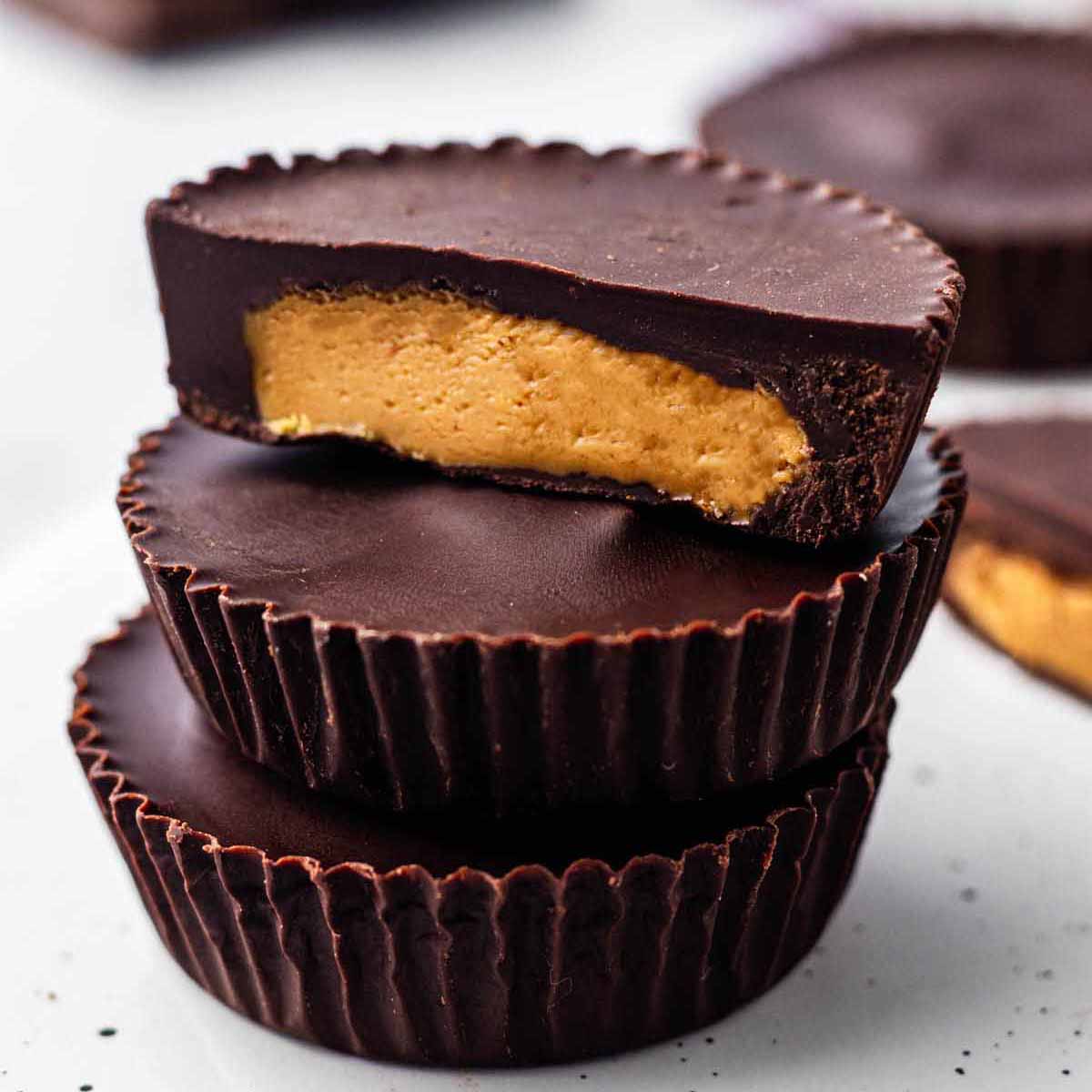 Three peanut butter cups stacked on a white surface. The top cup is cut in half to expose the creamy filling.