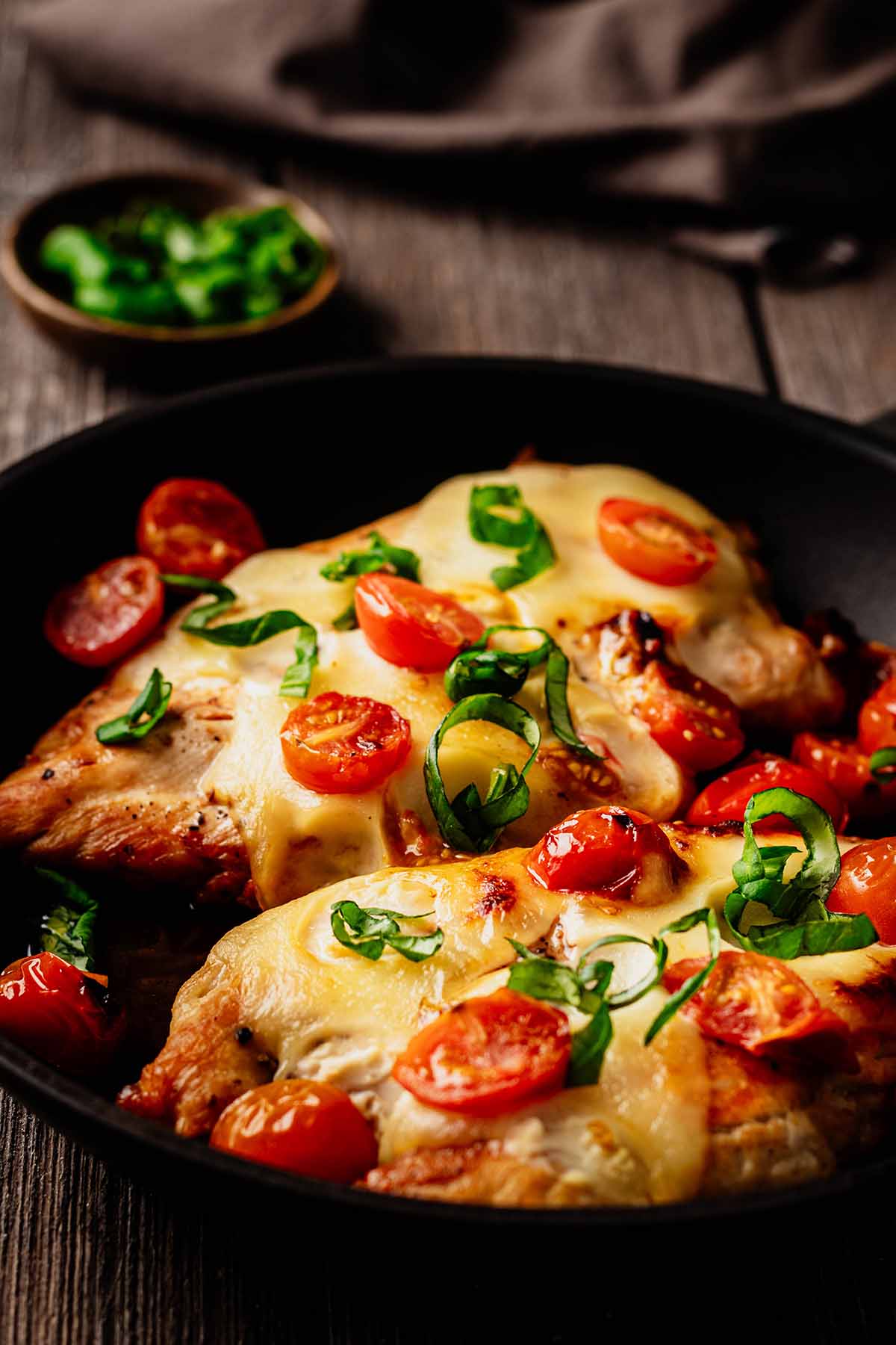 Close up of a two cooked chicken breasts in a skillet topped with melted mozzarella and cooked cherry tomatoes and garnished with chopped basil