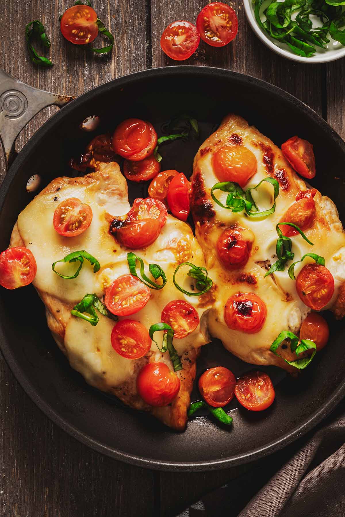 Overhead view of two cooked chicken breasts in a skillet topped with melted mozzarella and cooked cherry tomatoes and garnished with chopped basil