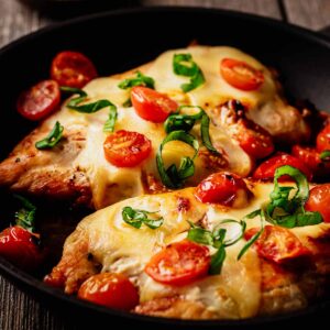 Close up of a two cooked chicken breasts in a skillet topped with melted mozzarella and cooked cherry tomatoes and garnished with chopped basil