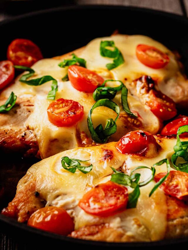 Close up of a two cooked chicken breasts in a skillet topped with melted mozzarella and cooked cherry tomatoes and garnished with chopped basil