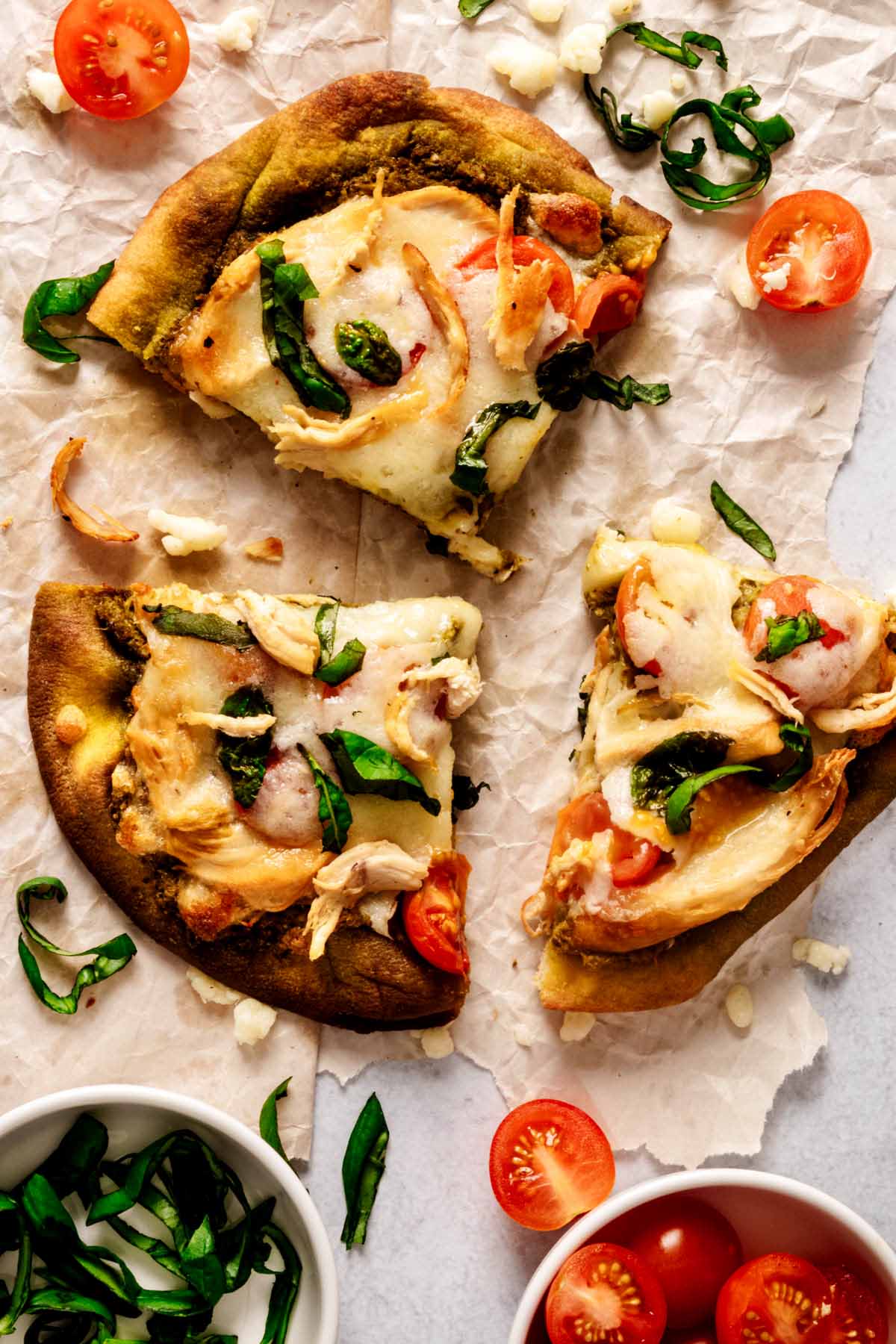 Overhead view of slices of chicken pesto flatbread on crumbled white paper with halved cherry tomatoes and chopped fresh basil