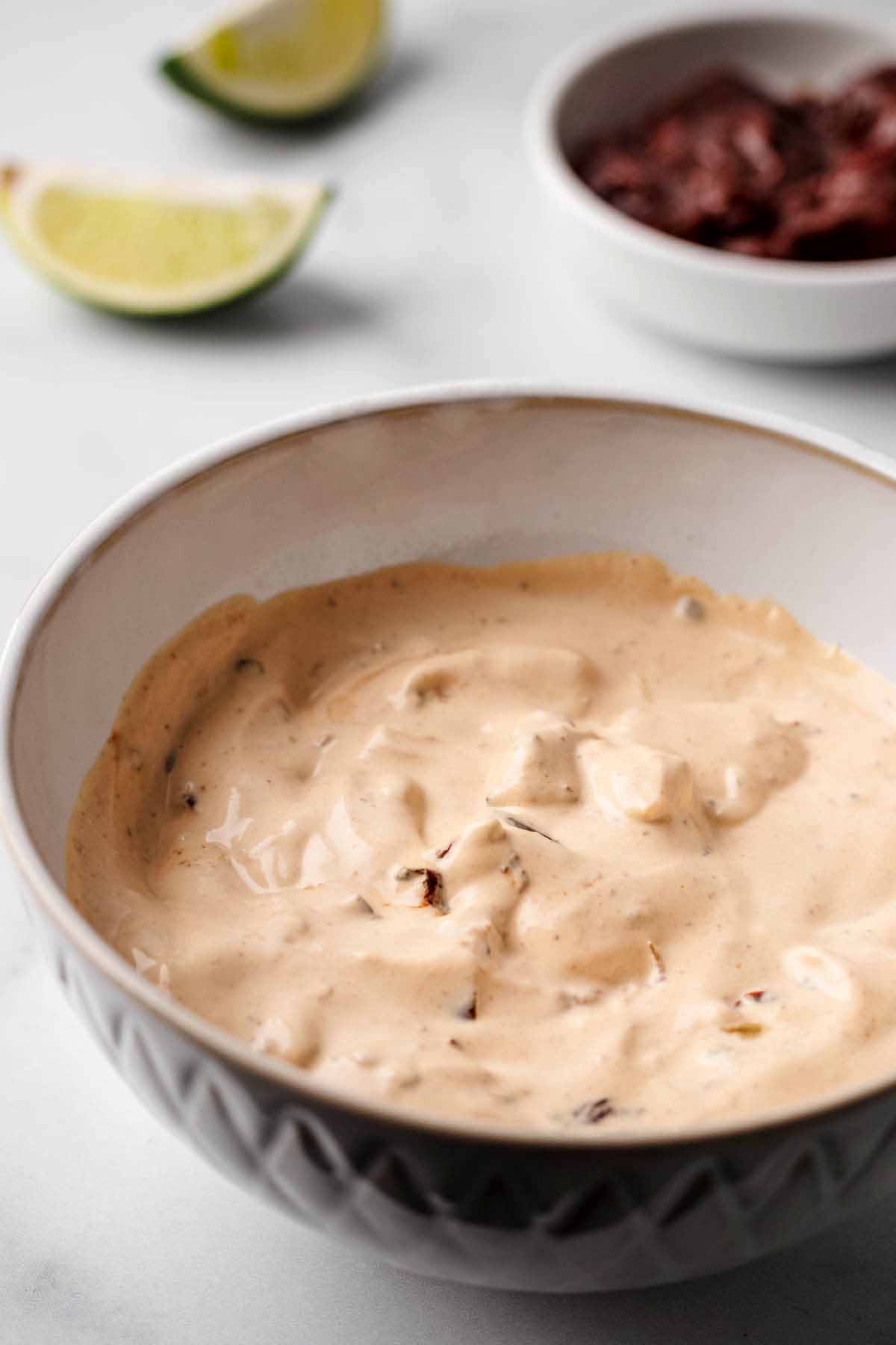 Close-up of a bowl of creamy chipotle sauce with visible chunks of chipotle peppers. In the background, lime wedges and a small bowl of chipotle peppers are slightly out of focus.