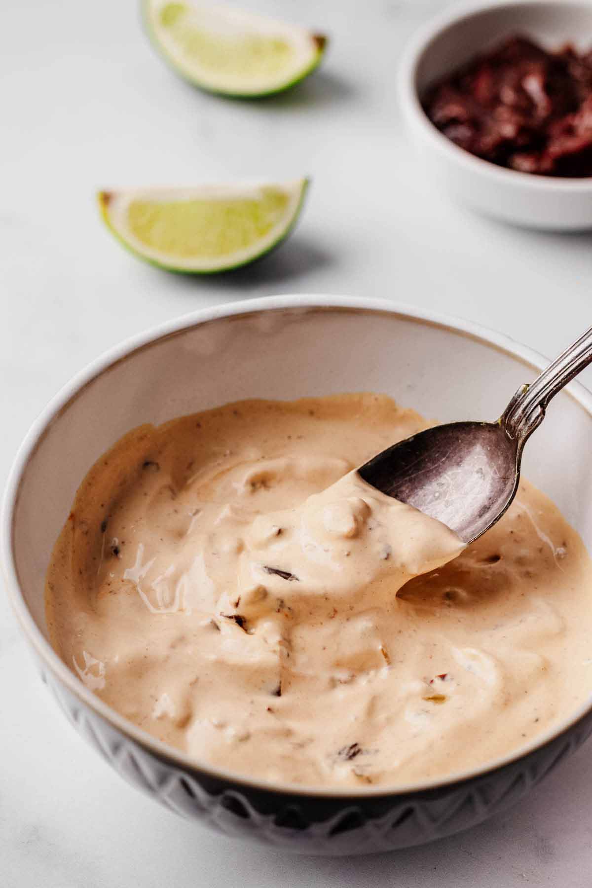 Close-up of a bowl filled with creamy chipotle sauce, with a spoon dipping into the sauce. Lime wedges and a small bowl of chipotle peppers are visible in the background.