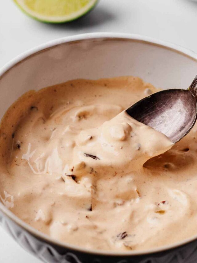 Close-up of a bowl of creamy chipotle sauce with visible chunks of chipotle peppers, being stirred with a spoon. A lime wedge is visible in the background.