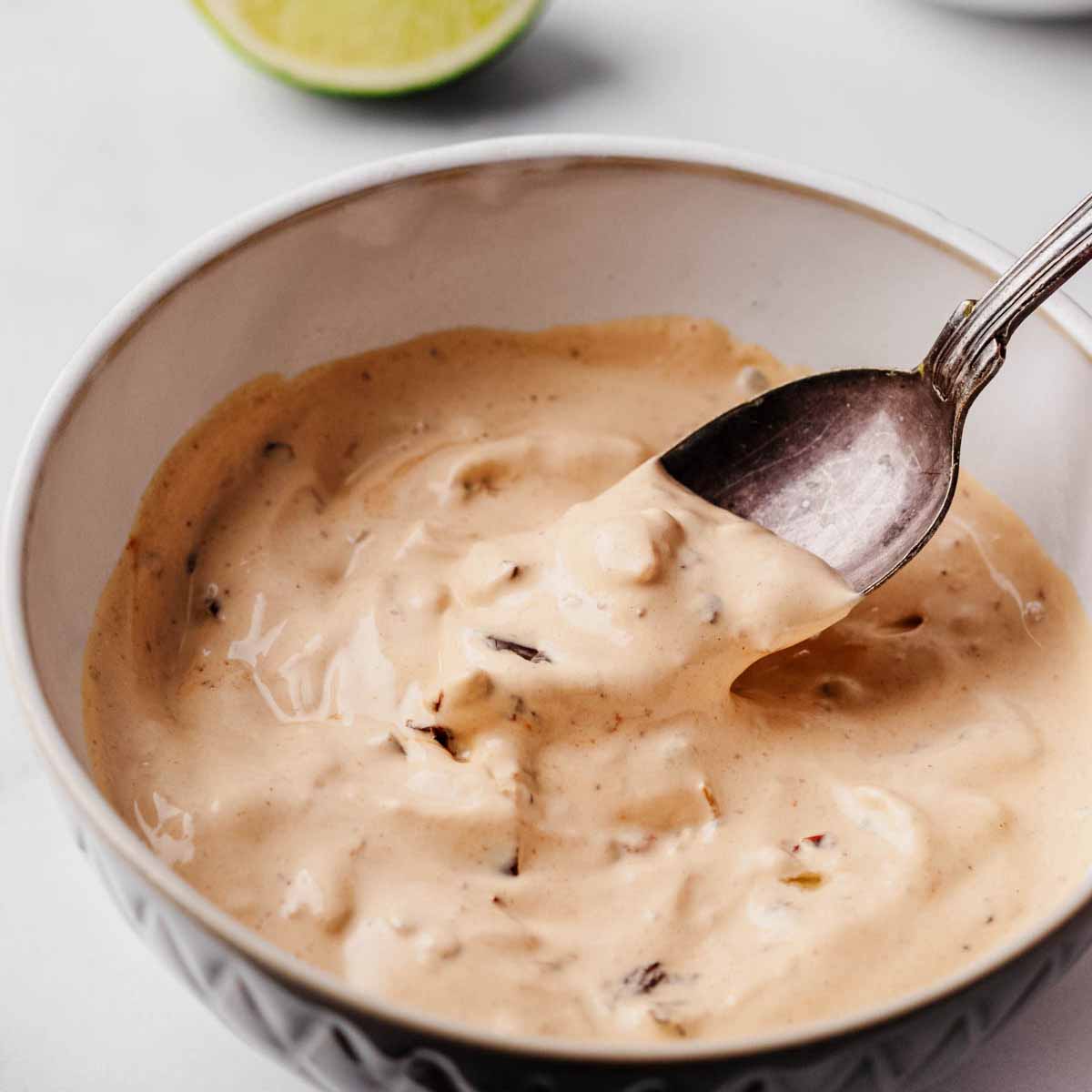 Close-up of a bowl of creamy chipotle sauce with visible chunks of chipotle peppers, being stirred with a spoon. A lime wedge is visible in the background.