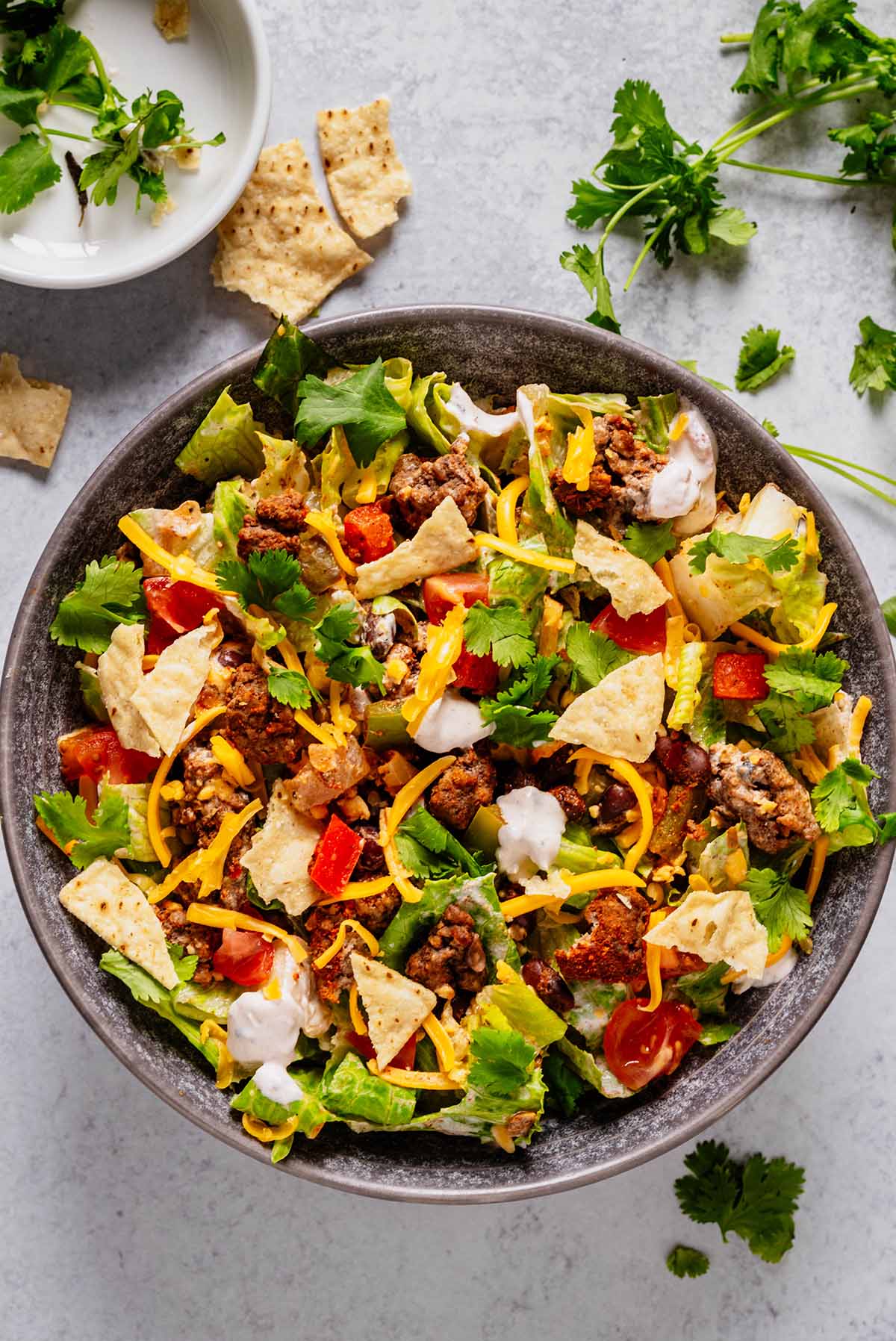 Top-down view of a ground beef taco salad with crisp romaine lettuce, seasoned ground beef, diced tomatoes, black beans, shredded cheddar cheese, crushed tortilla chips, and sour cream salsa dressing, garnished with fresh cilantro. A small bowl of cilantro and tortilla chips is visible in the upper left corner.