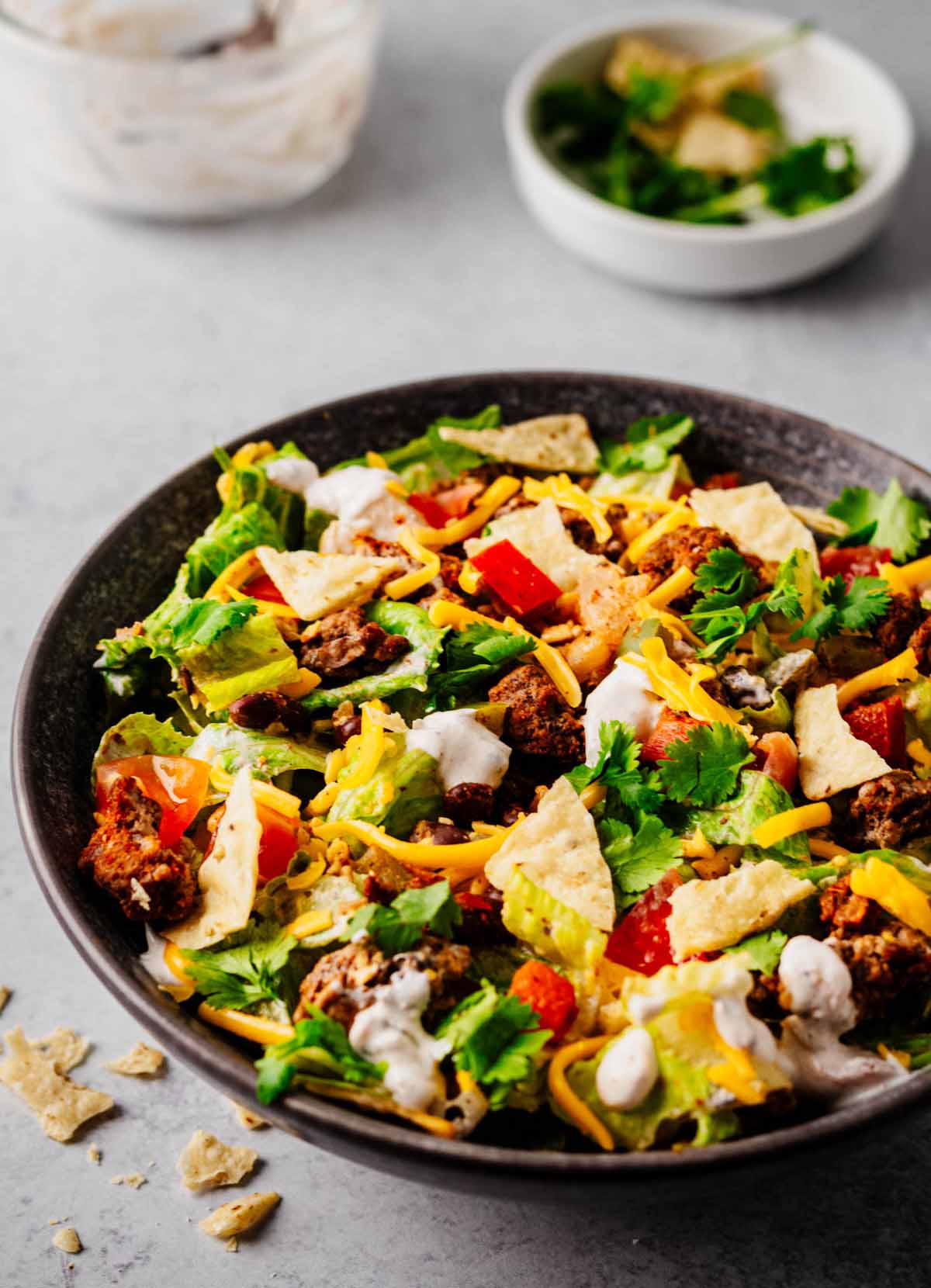 A bowl of ground beef taco salad featuring crisp romaine lettuce, seasoned ground beef, diced tomatoes, black beans, shredded cheddar cheese, crushed tortilla chips, and a sour cream salsa dressing, garnished with fresh cilantro. A small bowl of cilantro and tortilla chips is visible in the background.