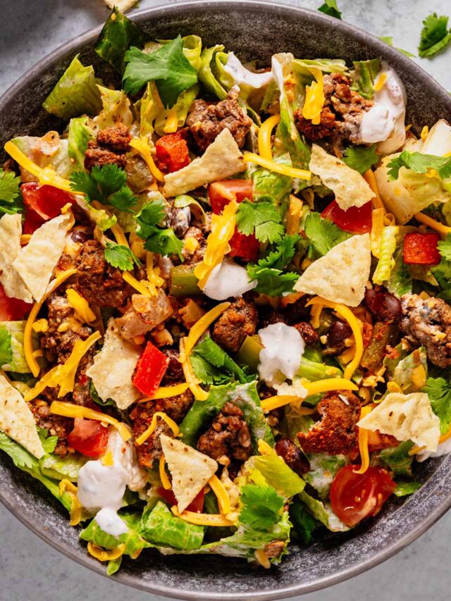 Top-down view of a colorful ground beef taco salad with romaine lettuce, seasoned ground beef, diced tomatoes, black beans, shredded cheddar cheese, crushed tortilla chips, sour cream salsa dressing, and fresh cilantro. Small scattered tortilla chips and cilantro garnish the surrounding surface.