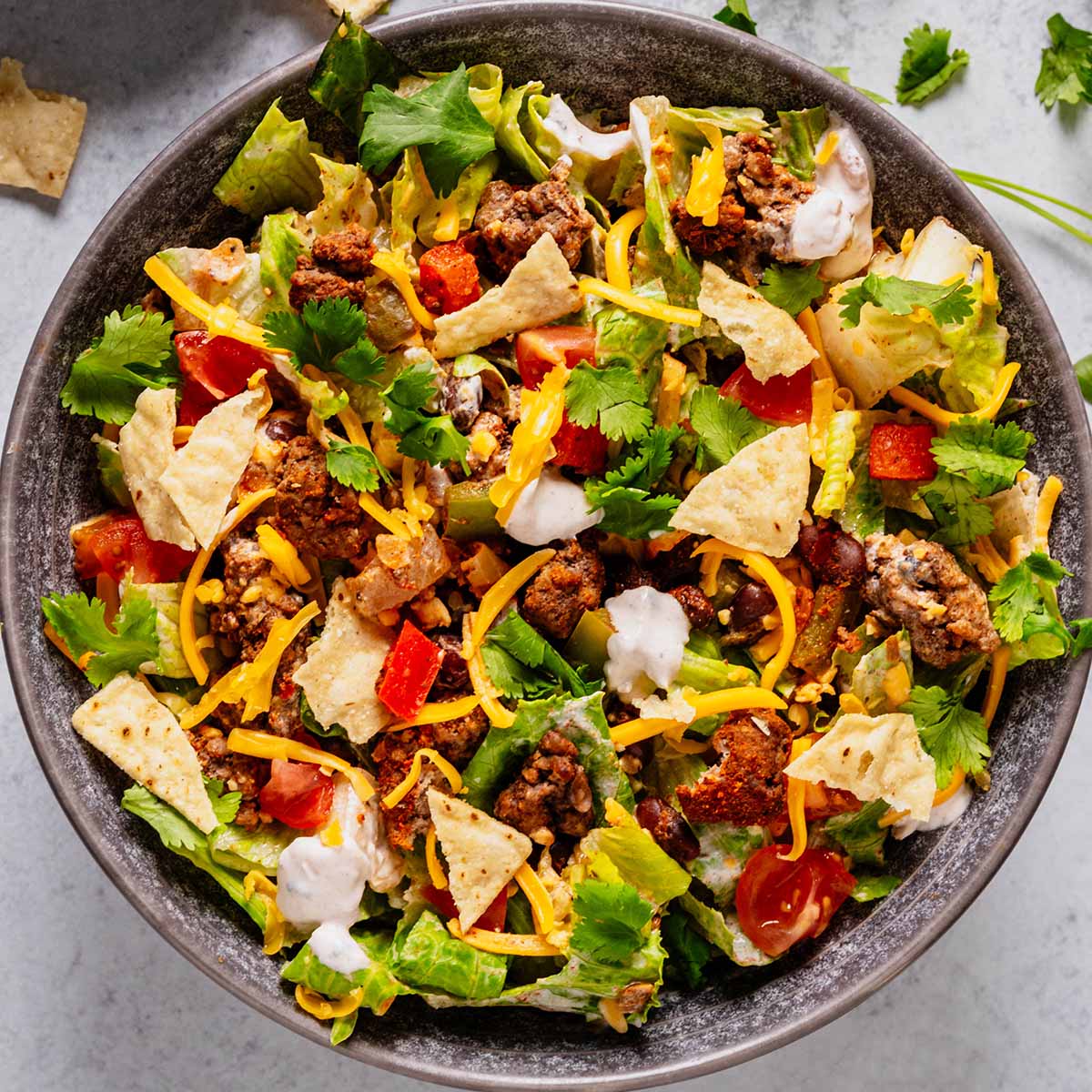 Top-down view of a colorful ground beef taco salad with romaine lettuce, seasoned ground beef, diced tomatoes, black beans, shredded cheddar cheese, crushed tortilla chips, sour cream salsa dressing, and fresh cilantro. Small scattered tortilla chips and cilantro garnish the surrounding surface.