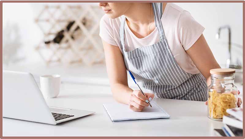 Young woman wearing an apron in the kitchen looking at a laptop and taking notes