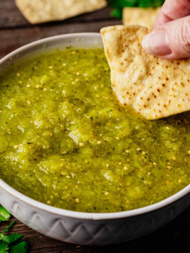 Tomatillo salsa in a small grey bowl with a tortilla chip being dipped into it.
