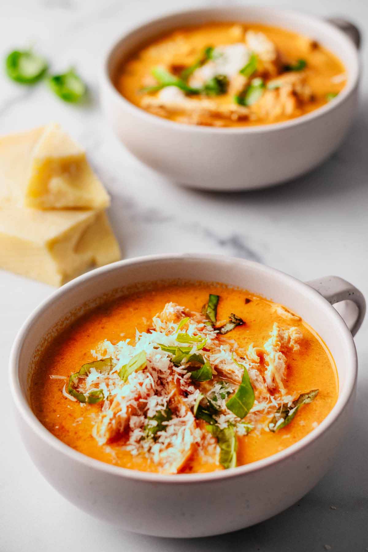 Tomato chicken soup topped with grated Parmesan and chopped basil in two white mug bowls