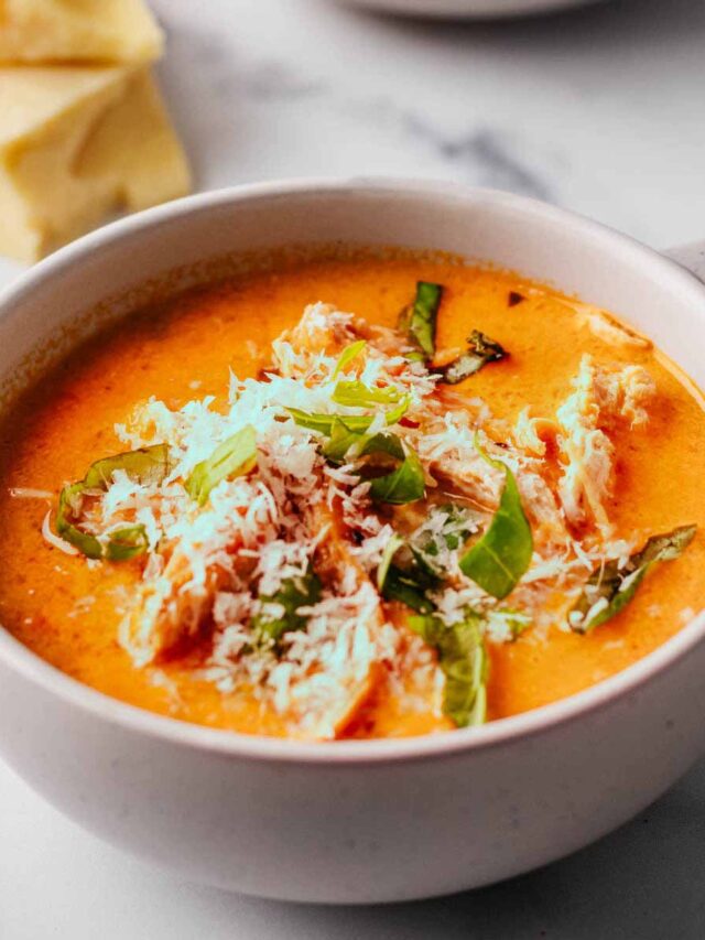 Tomato chicken soup topped with grated Parmesan and chopped basil in two white mug bowls