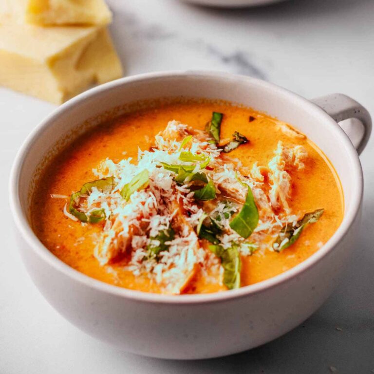 Tomato chicken soup topped with grated Parmesan and chopped basil in two white mug bowls