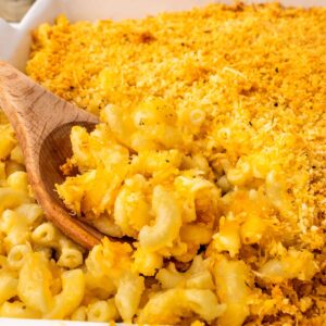 Close-up of creamy baked macaroni and cheese topped with a golden, crispy breadcrumb crust, with a wooden spoon scooping out a portion.