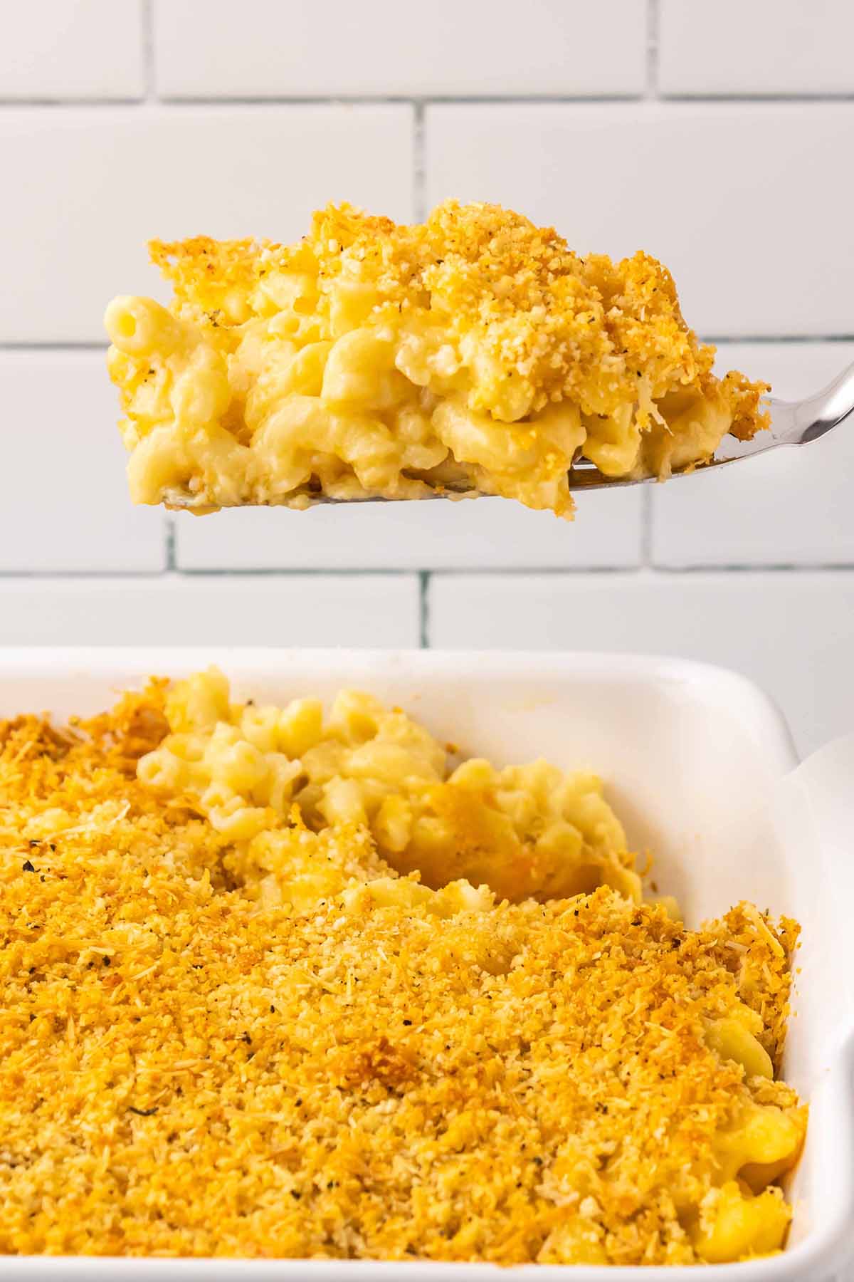 A slice of baked macaroni and cheese with a golden breadcrumb topping being lifted from a baking dish, showing its creamy interior.