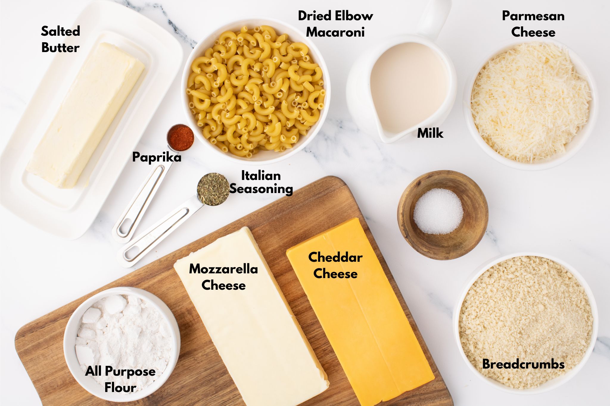 Overhead view of ingredients for baked macaroni and cheese, including salted butter, dried elbow macaroni, mozzarella cheese, cheddar cheese, parmesan cheese, breadcrumbs, all-purpose flour, milk, paprika, Italian seasoning, and salt, neatly arranged on a marble countertop.