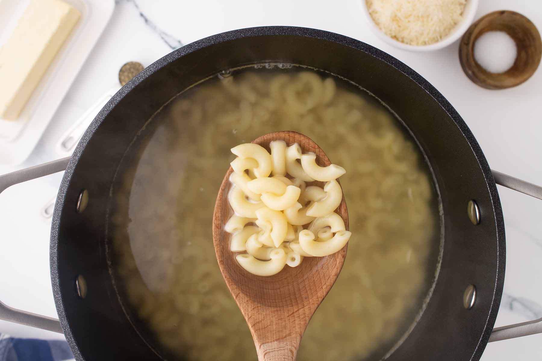 A wooden spoon holding cooked elbow macaroni over a pot of boiling water, preparing pasta for baked macaroni and cheese.