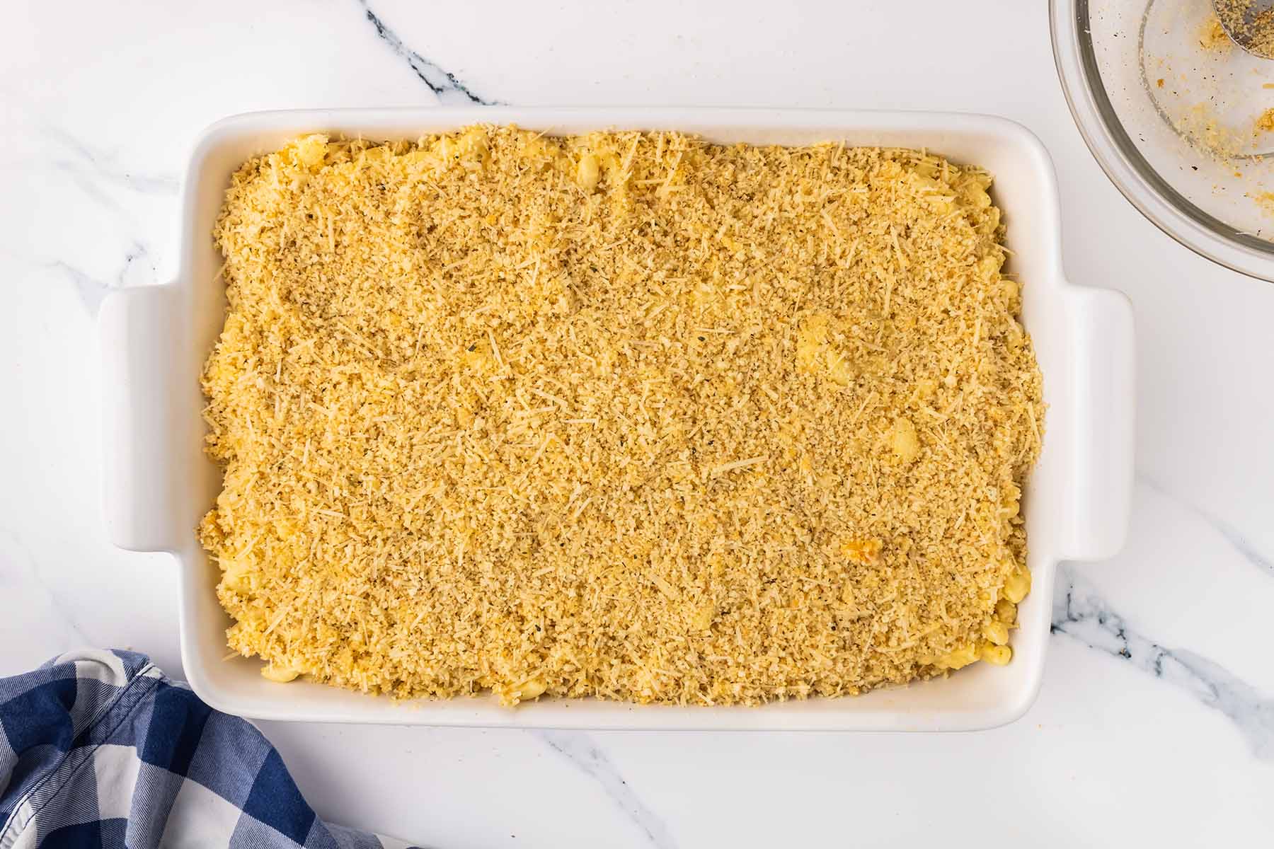 A baking dish filled with macaroni and cheese topped with an even layer of seasoned breadcrumbs and parmesan, ready to be baked.