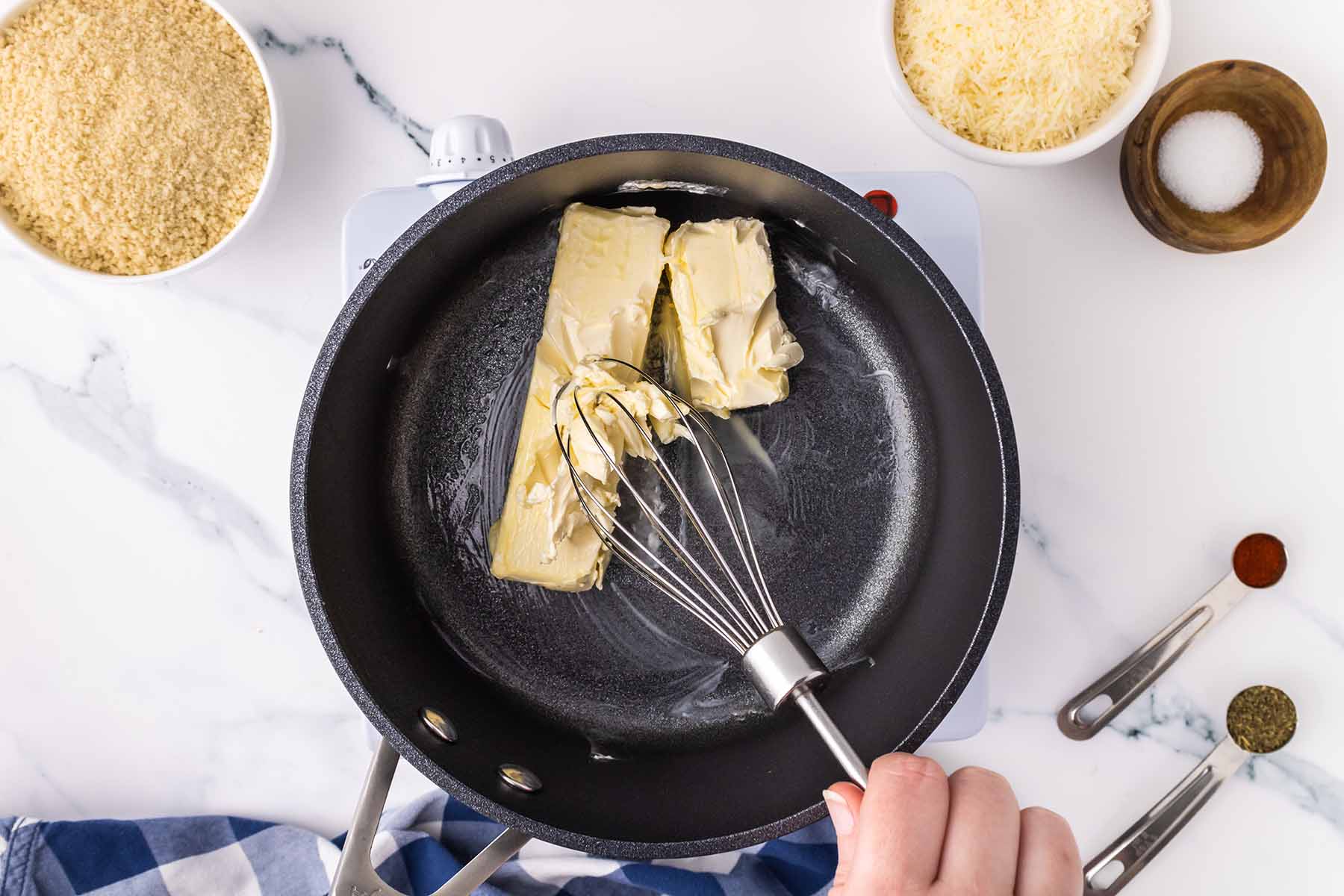 A person whisking butter as it melts in a black saucepan, preparing the roux for baked macaroni and cheese.